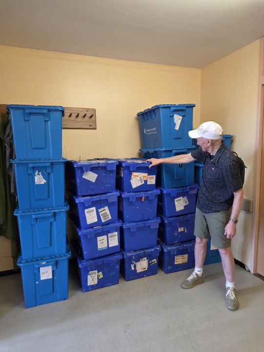 Steve Flaunty, the logistics team lead, in a white cap standing beside stacks of blue plastic storage boxes against a wall, some with shipping labels and one marked ARTHRO