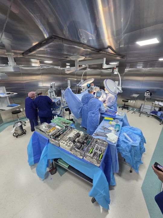 View from above of the surgical team around the operating table in a theatre with stainless-steel walls; trays of surgical instruments in the foreground and the patient covered by drapes