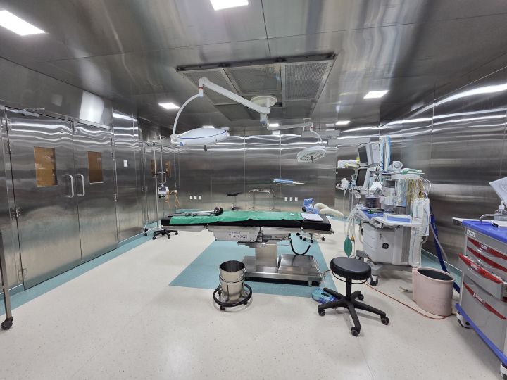 An empty operating theatre with stainless-steel walls, an operating table under a green drape, two large overhead surgical lights and an anaesthesia workstation
