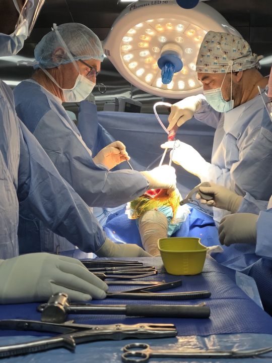 Dr John van Dalen (left) and Dr Tevita Tu'ungafasi (right), in masks and caps, working under the theatre light on a draped leg; the patient is not identifiable